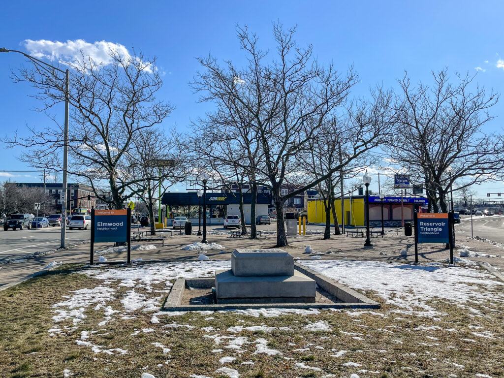 An empty plinth in the center of a small park between two busy streets. There is snow in the grass and the trees are bare.